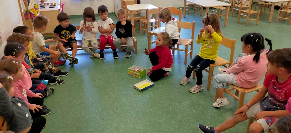 Bambini seduti in cerchio e al centro una bambina con una pigna in mano