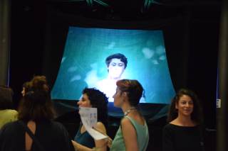 alcune persone riunite in una stanza dove viene trasmesso su uno schermo un\' immagine di un ragazzo