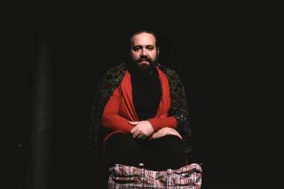 Foto dell\'attore seduto sul palco con maglia rossa e giacca nera sulle spalle.