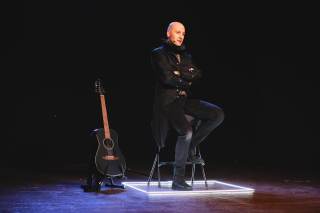 attore vestito di nero seduto su uno sgabello con accanto una chitarra