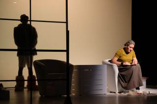 Foto degli attori sul palco. Un\'attrice &egrave; seduta su una poltrona mentre l\'altro si trova alle sue spalle non illuminato.