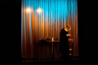 Foto dell\'attrice sul palco. L\'attrice, davanti al pubblico, tiene un panno in una mano e una sigaretta nell\'altra.