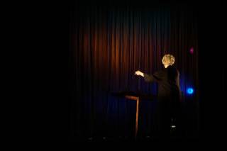 Foto dell\'attrice sul palco. L\'attrice gesticola sul palco davanti al tavolo.