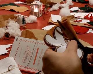 Foto di uno dei tavoli del laboratorio pieno di fogli di carta, fili e tessuti. Un partecipante sta ritagliando.