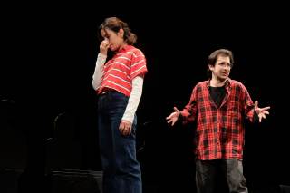 Foto dello spettacolo. Uno dei due attori parla, l\'altra tiene una mano sul naso.