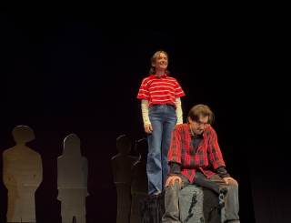 Foto dello spettacolo. Un attore &egrave; seduto, l\'altra attrice &egrave; in piedi e sorride alle sue spalle.