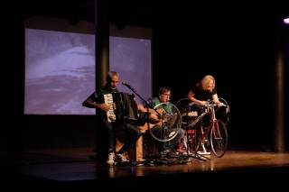 Foto dello spettacolo. L\'attrice &egrave; a cavallo di una bici, con gli altri attori accanto.