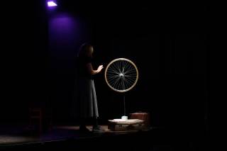 Foto dello spettacolo. L\'attrice sta accanto ad una ruota di una bici illuminata dalla luce.