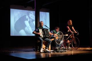 Foto dello spettacolo. L\'attrice &egrave; a cavallo di una bici, con gli altri attori accanto. Sul fondo, una foto di Alfonsina Strada.