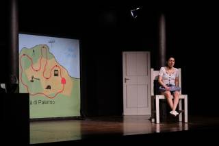 Foto dello spettacolo. Un\'attrice &egrave; seduta su una sedia con aria confusa. Sul fondo, una mappa della citt&agrave; di Palermo.