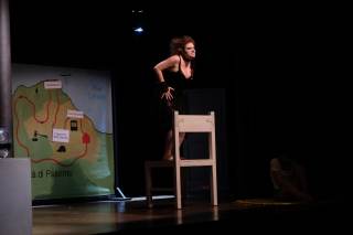 Foto dello spettacolo. Un\'attrice &egrave; in piedi su una sedia con fare aggressivo.