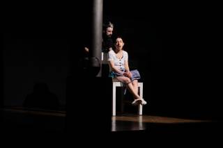 Foto dello spettacolo. Un\'attrice &egrave; a sedere e parla, dietro di lei un altro attore si avvicina con fare minaccioso.